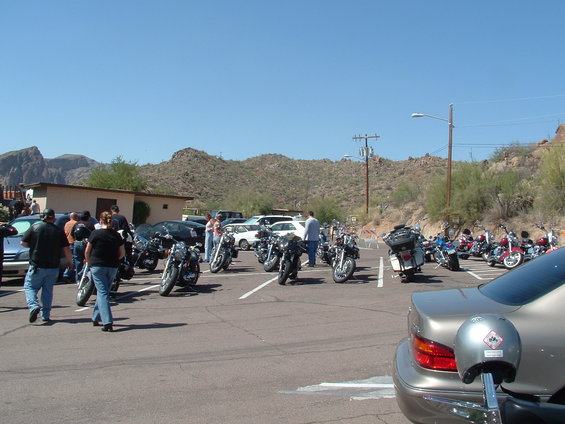 Motorcycles and cars parked in a dusty lot with mountains in the background