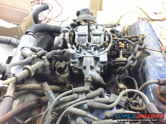 Engine block with carburetor, spark plug wires, and hoses. A chain is wrapped around part of the engine