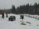 1979 Ford Bronco - Two SUVs navigate a snowy landscape, with a wooden fence and dense pine forest in the background.