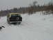 1979 Ford Bronco - A 1979 Ford Bronco with a tan top drives through a snowy landscape with trees in the background.