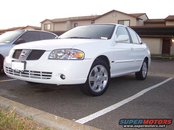 White Nissan Sentra parked in a lot, another car partially visible to the left