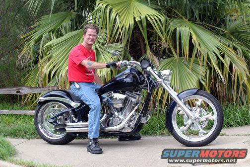Man in red shirt and jeans sits on a black custom motorcycle with chrome accents. Palm trees and a wooden fence are in the background. "SUPERMOTORS www.supermotors.net" logo is in the lower right