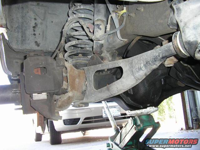 Rear suspension components including coil spring, brake caliper, differential housing, and control arm. A floor jack supports the differential
