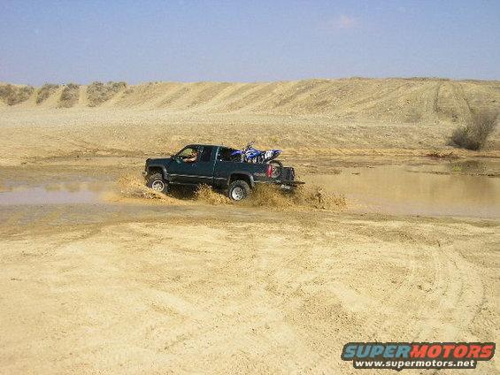 Green pickup truck with a dirt bike in the bed driving through mud, side view