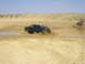 1994 Ford Bronco - A green pickup truck with a dirt bike in its bed splashes through a muddy puddle in a dry, sandy landscape.