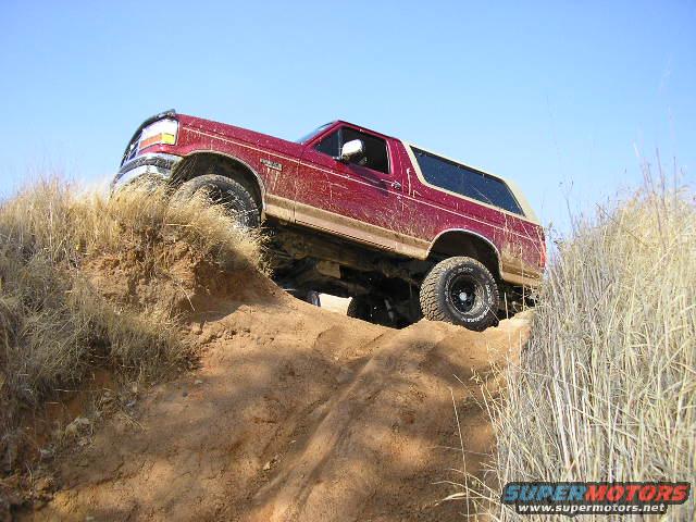 Red Ford Bronco climbing a dirt mound, tires kicking up dust, clear blue sky above