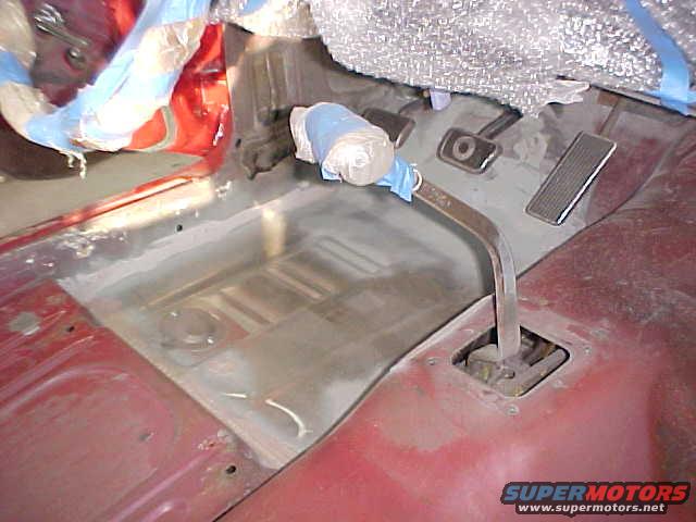Car interior floor pan with pedals and a gear shifter. Some parts are covered in plastic wrap
