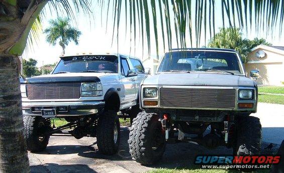 Two lifted pickup trucks parked side-by-side. The one on the left has a visor with 'D-ROCK' and a license plate that says 'D-ROCK'. The one on the right is a different model