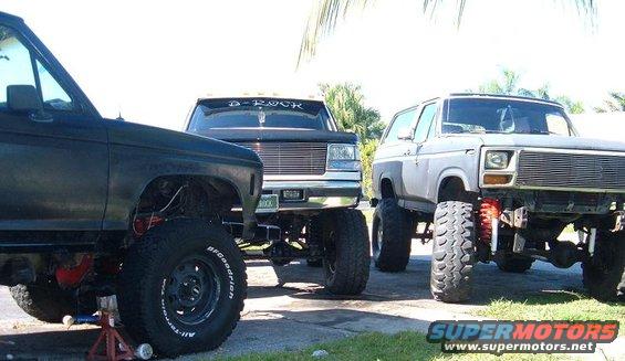 Three lifted trucks parked side-by-side, front view. The truck on the right is a 1978 Ford Bronco