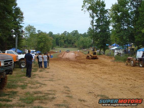 Mud bog event with multiple off-road vehicles, a bulldozer, and spectators gathered around a dirt track