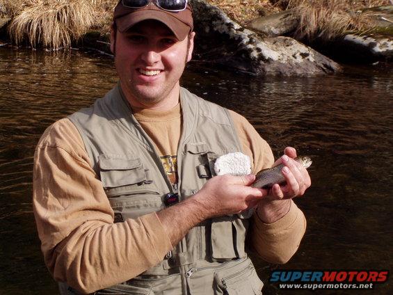 Man in fishing vest holding a small trout by a river