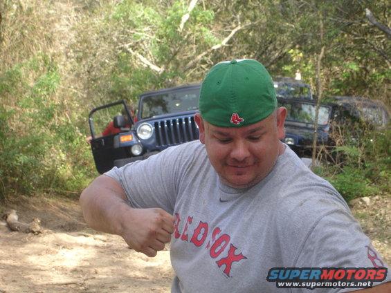 Guy wearing a green Red Sox hat and gray t-shirt, with a blue Jeep behind him on a dirt path