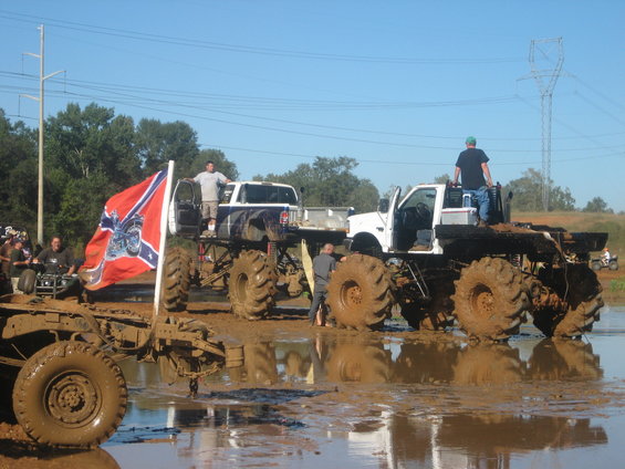 Muddy trucks with large tires in a bog, one with a Confederate flag