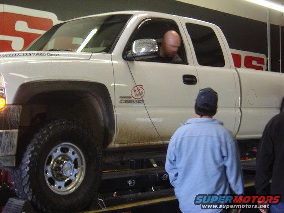 White pickup truck with "6.6L Duramax Turbo Diesel" badging on a dynamometer, driver visible inside