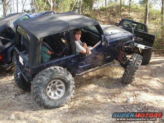 Modified dark blue Jeep Wrangler with large tires and a soft top, off-roading with another vehicle behind it