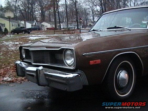 Brown sedan front corner with chrome grille and bumper. Two trucks in background