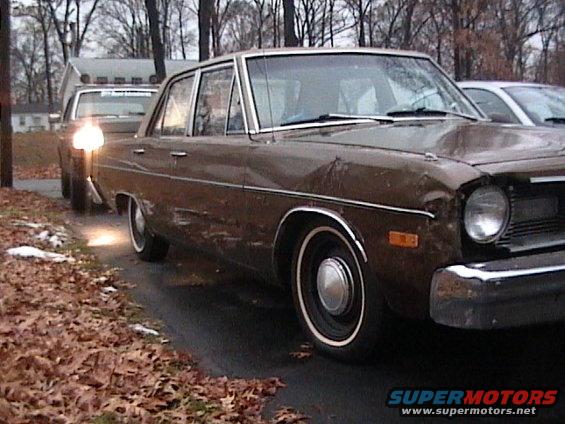 Brown sedan parked on a gravel driveway with fallen leaves, another car behind it with its headlights on