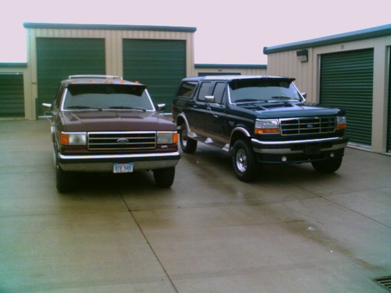 Two Ford trucks parked in front of storage units. The truck on the left is maroon, and the truck on the right is dark blue. Both have sun visors