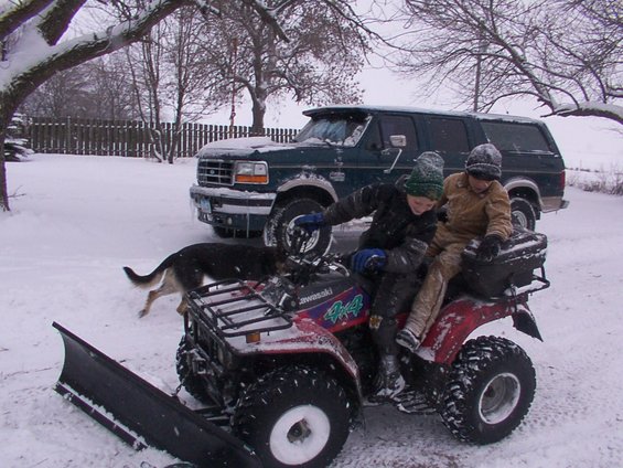 Green Ford F-Series truck in snow, front view. Two children on a red ATV in foreground