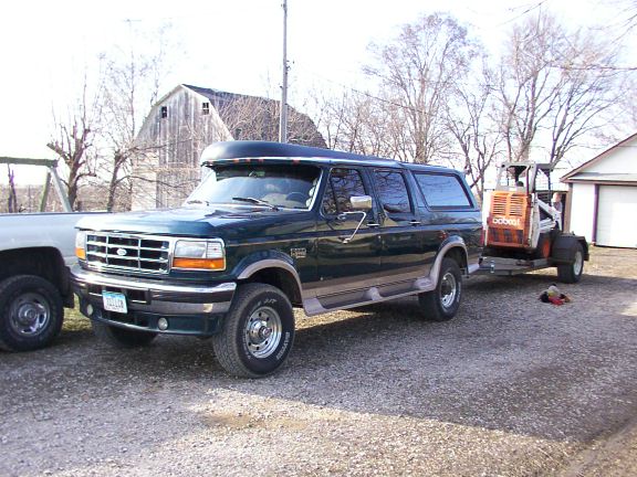 1995 Ford C-350 Centurion Conversion van, dark green, towing a Bobcat skid steer loader. Another truck is partially visible on the left