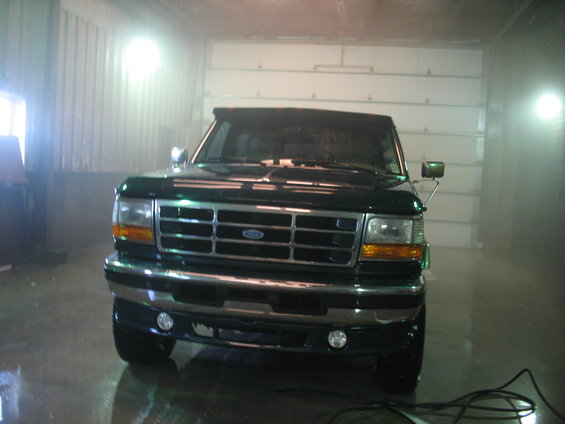 Front view of a dark green Ford F-Series truck in a garage. It has a chrome grille and fog lights