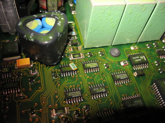 Close-up of a green circuit board with a black triangular component containing blue and yellow capacitors, and several integrated circuits