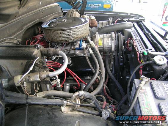 Engine bay with V8 engine, air filter, battery, and hoses. A blue Chevy Van is visible in the background