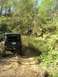 1989 Ford Bronco - An off-road vehicle navigates a muddy trail surrounded by dense foliage, with another vehicle visible in the distance.