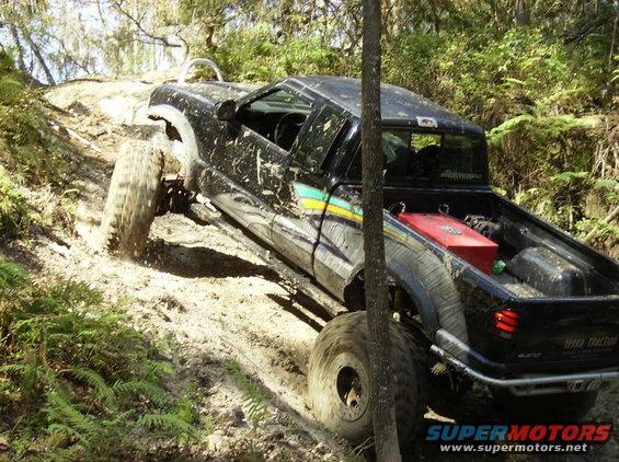 Black pickup truck with large tires climbing a steep, muddy, wooded trail. It has a red fuel tank in the bed