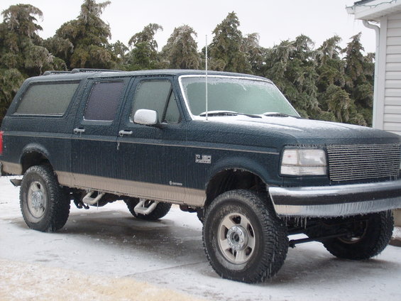 Dark green SUV with tan trim covered in ice, parked on a snowy surface with icy trees in the background