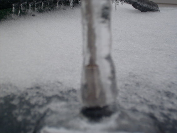 Close-up of an icicle hanging over snow with a dark, blurred object in the background