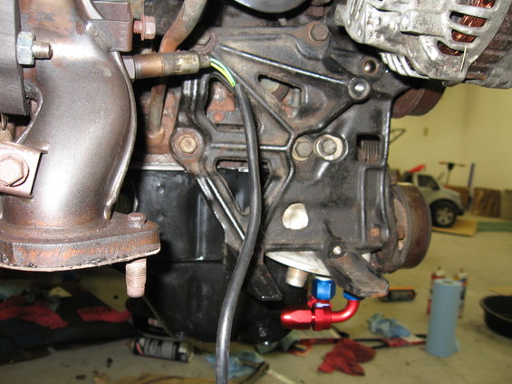 Engine block with exhaust manifold, alternator, and oil lines. A silver car is visible in the background