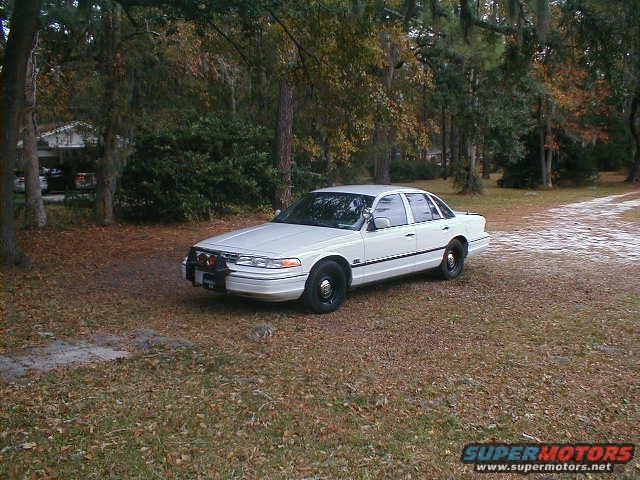 White sedan parked on a grassy area with trees in the background. The car has a push bumper with lights