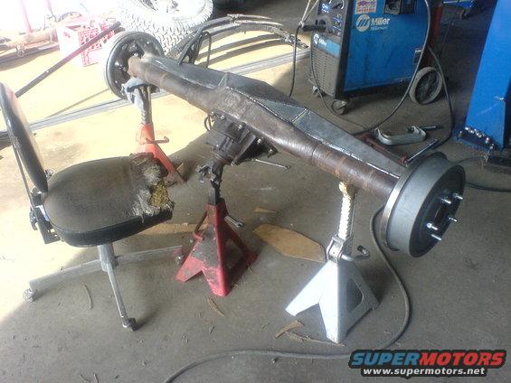 Rear axle assembly on jack stands with a torn office chair nearby and a welding machine in the background