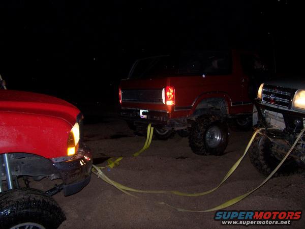 Red pickup truck, red Bronco, and silver SUV are hooked up with tow straps at night