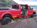 1991 Ford Bronco - A red pickup truck is stuck in soft sand with its doors open, showing the interior and the wheels buried.