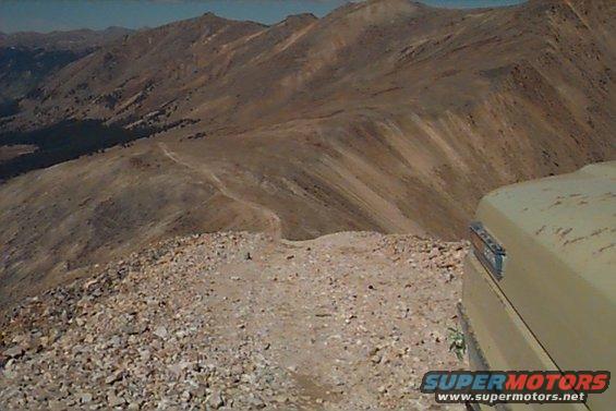 Mountain landscape with a dirt road and the corner of a tan vehicle visible on the right. Watermark reads SUPERMOTORS.NET