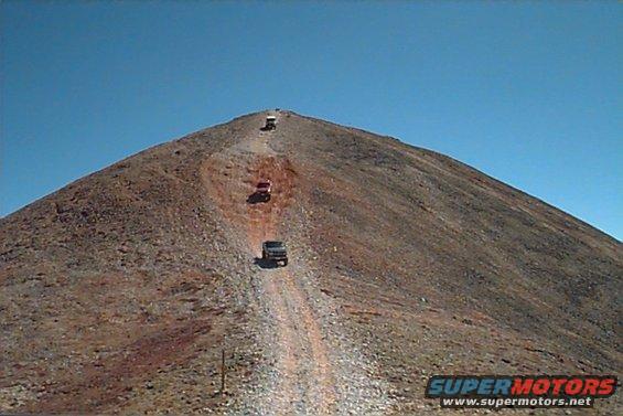 Several SUVs driving up a steep, rocky mountain trail under a clear blue sky. The lead vehicle is dark, followed by a red one, then a white one