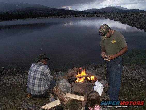 Two men and a child by a campfire next to a lake with mountains in the background