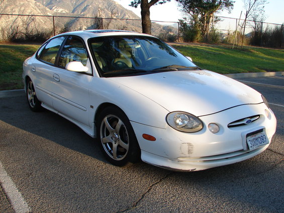White sedan with aftermarket wheels and front bumper damage, parked on asphalt