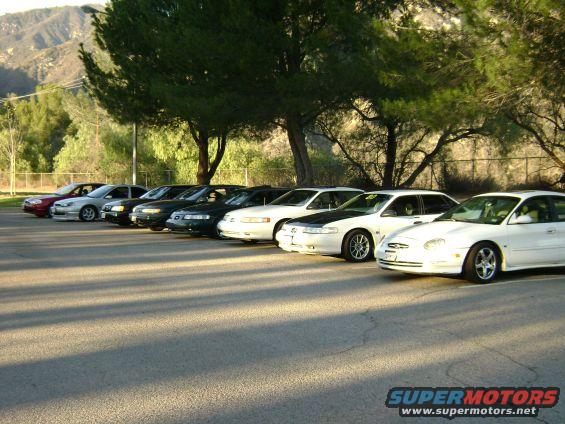 Several sedans parked in a row, some white, some dark. Hills and trees in background