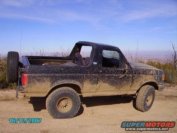 Mud-covered black 1990 Ford Bronco parked on a dirt road with a spare tire mounted on the rear. The vehicle is seen from the side