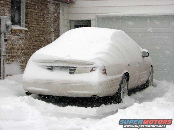 Rear view of a white 2003 Ford Crown Victoria covered in snow, parked next to a garage