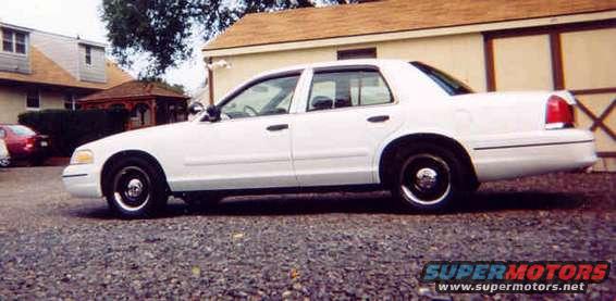 White sedan parked on gravel lot, side view. Red car in background