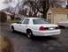 1999 Ford Crown Victoria - A white 1999 Ford Crown Victoria is parked in front of a house and shed, with another car covered in a tarp nearby.