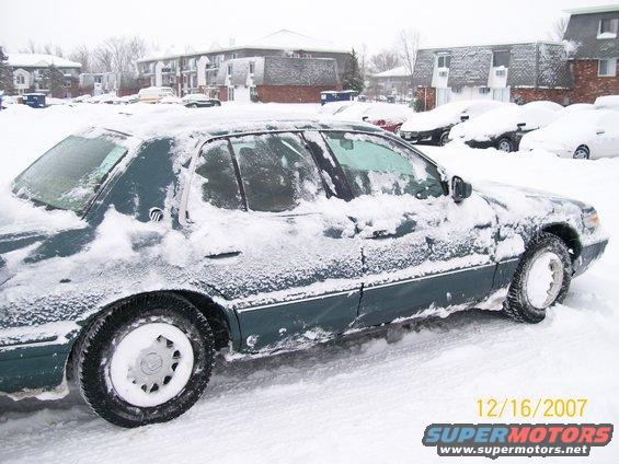 Dark green sedan covered in snow, side view, with a SuperMotors watermark