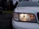 1999 Ford Crown Victoria - A white sedan's front corner is shown, with the headlight on, and another vehicle in the background.