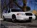 1999 Ford Crown Victoria - A white 1999 Ford Crown Victoria Police Interceptor is shown from a 3q-front angle, with text indicating the year and model visible at the…