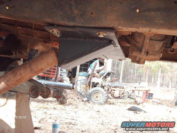 Custom bracket bolted to underside of vehicle frame, near drivetrain components. Tractor and ATV visible in background