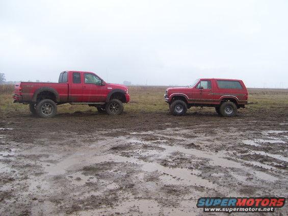 1986 Ford Bronco and a red pickup truck parked side-by-side in a muddy field